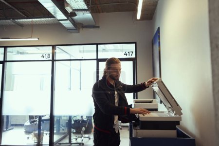 Foto de Hombre joven en ropa de mezclilla casual está utilizando una fotocopiadora en un espacio de coworking, la habitación es limpia y espaciosa - Imagen libre de derechos