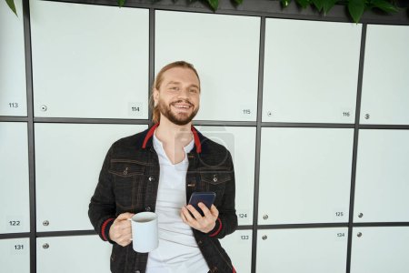 Téléchargez les photos : Homme heureux avec une tasse de café se tient près des casiers dans un espace de coworking, un homme en vêtements décontractés - en image libre de droit