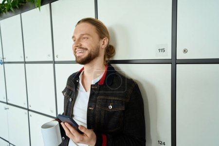 Photo for Smiling male with a cup of coffee and phone stands by the lockers in coworking space, male in casual clothes - Royalty Free Image
