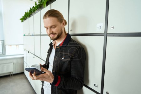 Photo for Guy in casual clothes is standing at the lockers in a coworking space, a male is texting on the phone - Royalty Free Image