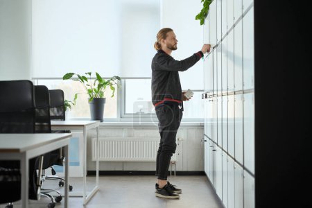 Foto de Barbudo cierra la puerta de un casillero en un espacio de coworking, el macho tiene una taza en sus manos - Imagen libre de derechos