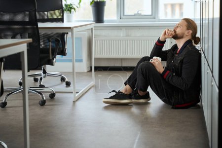 Photo for Frustrated employee with a cup of coffee sits on the floor near lockers in a coworking space - Royalty Free Image