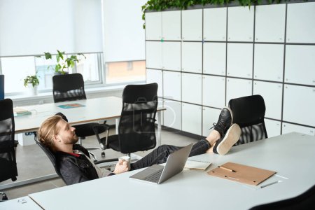 Téléchargez les photos : Jeune employé est situé dans un espace de coworking avec un ordinateur portable et café, il a mis ses pieds sur la table - en image libre de droit
