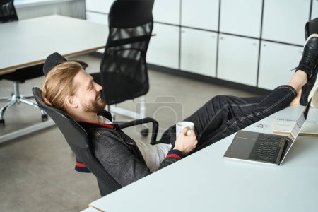 Photo for Young employee communicates by laptop and drinks coffee, he put his feet on the table - Royalty Free Image
