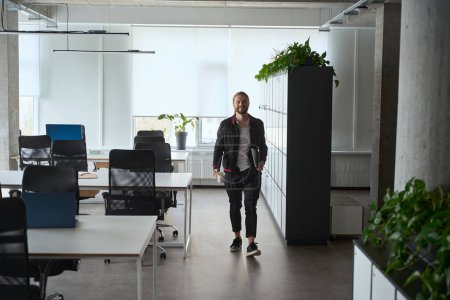 Foto de Hombre alegre en ropa casual de mezclilla camina alrededor del espacio de coworking, tiene una computadora portátil en sus manos - Imagen libre de derechos