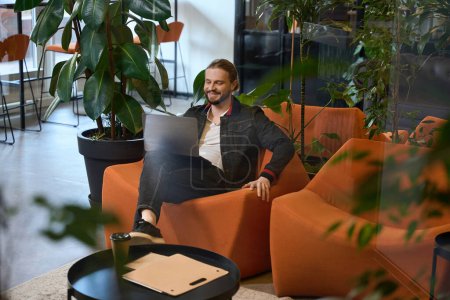 Photo for Smiling male comfortably settled down with a laptop in the lounge area, he communicates on the laptop - Royalty Free Image