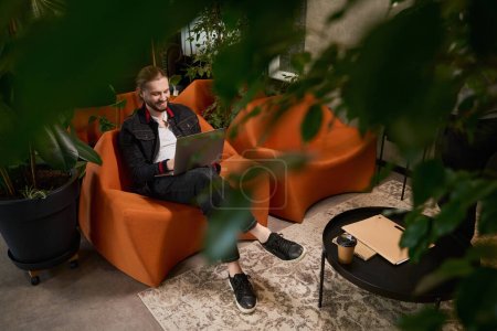 Foto de Guy se encuentra en silla acogedora en la sala de estar con ordenador portátil, junto a una carpeta con documentos y café - Imagen libre de derechos