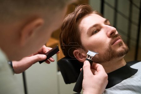 Foto de Peluquero macho trabaja con un cliente en una barbería, el maestro utiliza un recortador de barba especial - Imagen libre de derechos