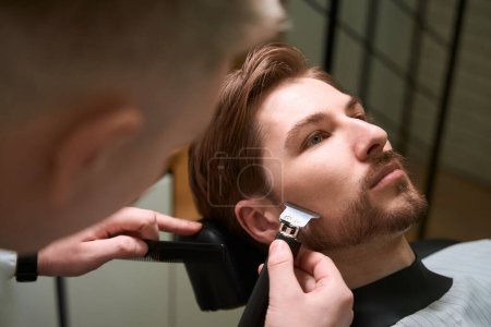 Foto de Maestro peluquero trabaja con un cliente joven en una barbería, un especialista utiliza un cortador de barba - Imagen libre de derechos
