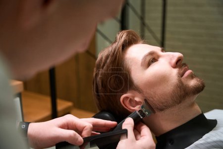 Foto de Maestro corta la barba de un joven cliente en una barbería, un especialista utiliza una podadora de barba - Imagen libre de derechos