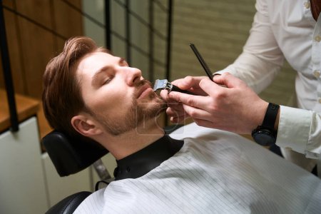 Téléchargez les photos : Homme coupe une barbe à un jeune client dans un salon de coiffure, un spécialiste utilise des outils spéciaux - en image libre de droit