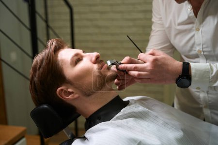 Foto de Maestro alinea la línea de bigote a un cliente joven en una barbería, el especialista utiliza herramientas especiales - Imagen libre de derechos