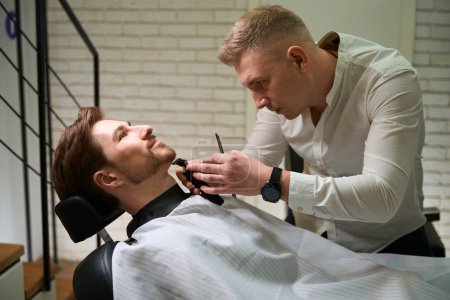 Photo for Young barber shaves beard of handsome man in barbershop, specialist uses beard trimmer - Royalty Free Image