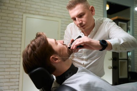 Téléchargez les photos : Jeune homme assis dans une chaise spéciale dans un salon de coiffure, le maître travaille avec sa barbe - en image libre de droit