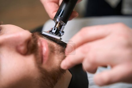 Téléchargez les photos : Maître coiffeur au travail s'occupant d'un client barbe et moustache, un professionnel utilise un outil spécial - en image libre de droit