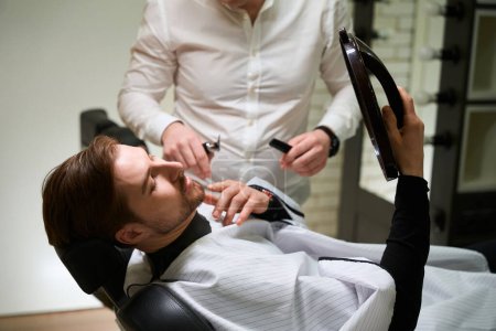 Foto de Elegante hombre se sienta en una silla en la barbería y se mira en el espejo, al lado del maestro barbero con herramientas especiales - Imagen libre de derechos