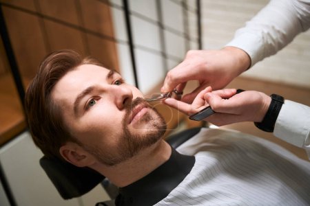 Photo for Barber at work caring for a clients mustache, the master uses special scissors - Royalty Free Image