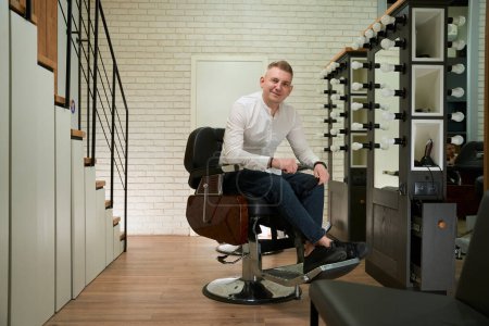 Photo for Man barber sits in a barber chair at his workplace, he has professional tools in his hands - Royalty Free Image