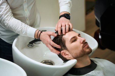 Photo for Hairdresser in the barbershop washes the hair of young man, the client is located in special chair by the sink - Royalty Free Image
