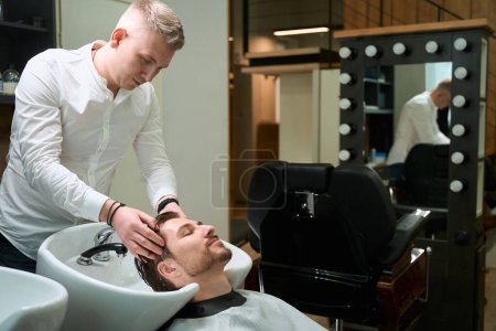 Téléchargez les photos : Jeune homme aime le processus de lavage de ses cheveux dans un salon de coiffure, un coiffeur travaille sur un lieu de travail bien équipé - en image libre de droit