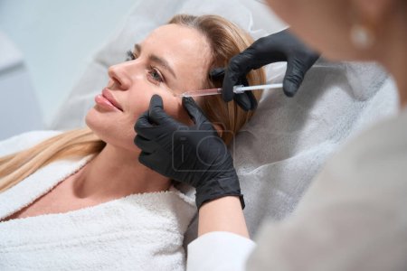Foto de Mujer rubia recibe una inyección rejuvenecedora en sus mejillas, un esteticista utiliza una aguja delgada - Imagen libre de derechos