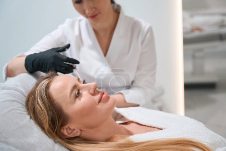 Photo for Doctor in uniform makes anti-aging injections around the eyes of a client, an esthetician uses a thin needle - Royalty Free Image