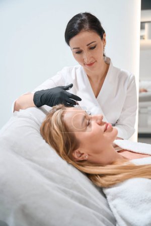 Foto de Esteticista en uniforme realiza un procedimiento de inyección alrededor de los ojos de una mujer rubia, un esteticista utiliza una aguja delgada - Imagen libre de derechos