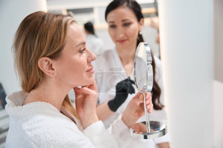 Téléchargez les photos : Charmante femme se regarde dans le miroir lors d'une consultation avec un médecin esthéticienne, spécialiste en uniforme - en image libre de droit