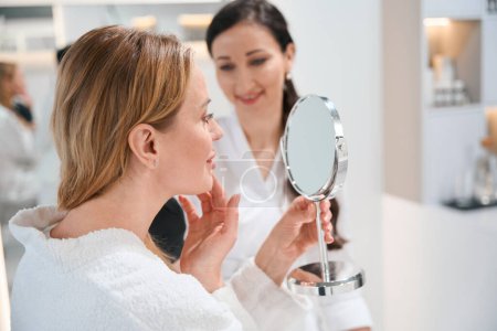 Téléchargez les photos : Brunette cosmétologue consulte une cliente blonde dans une clinique de cosmétologie, les regards féminins dans le miroir - en image libre de droit
