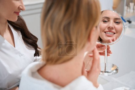 Photo for Smiling female blonde looks in mirror at consultation with an esthetician, she is pleased with the reflection in the mirror - Royalty Free Image