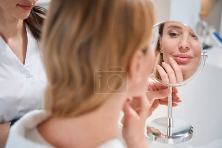 Photo for Client of a cosmetology clinic examines her face in a mirror at a consultation with an esthetician - Royalty Free Image