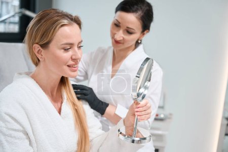 Patientin einer kosmetologischen Klinik untersucht ihr Gesicht in einem Spiegel bei einer Konsultation beim Spezialisten
