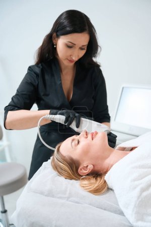 Estética femenina realiza un procedimiento de rejuvenecimiento de hardware, el cliente se encuentra cómodamente ubicado en un sofá de cosmetología
