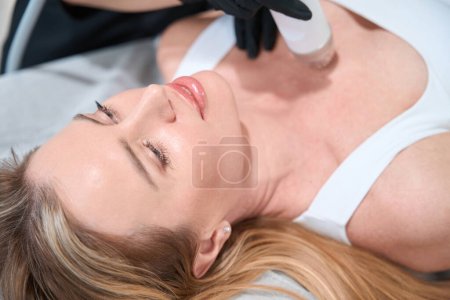 Foto de Cosmetóloga femenina realiza procedimiento de elevación de RF en el área decoleta, el cliente se encuentra en un sofá de cosmetología - Imagen libre de derechos