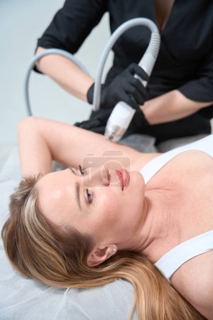 Téléchargez les photos : Esthéticienne médecin effectue une procédure de levage RF efficace sur la région de l'épaule du client, la femme se trouve sur le canapé cosmétique - en image libre de droit