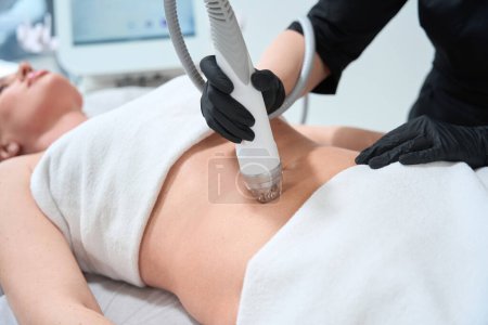 Mujer joven se encuentra en un sofá de cosmetología, un esteticista realiza un procedimiento de elevación de RF para su abdomen