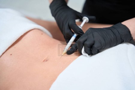 Foto de Empleado de una clínica de cosmetología hace inyecciones en el estómago de un cliente, un esteticista trabaja en guantes de protección - Imagen libre de derechos