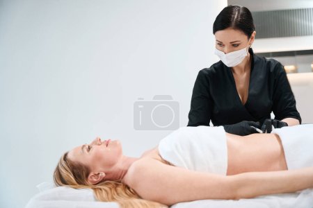 Foto de Mujer con un uniforme negro hace inyecciones en el abdomen de un cliente, un esteticista trabaja en guantes de protección - Imagen libre de derechos