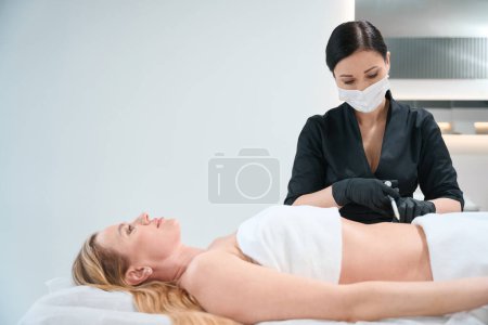 Mujer en uniforme negro hace inyecciones en el abdomen del cliente, un esteticista trabaja en guantes de protección y una máscara