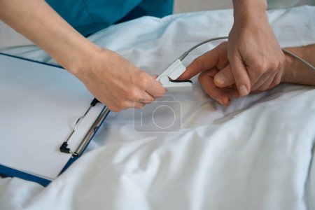 Foto de Primer plano del médico colocando oxímetro de pulso en el dedo del paciente masculino que yace en la cama en la clínica - Imagen libre de derechos