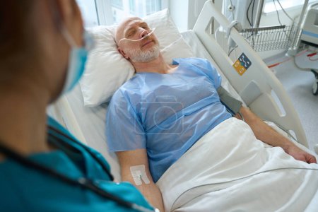 Téléchargez les photos : Médecin féminin parlant à un patient de sexe masculin allongé dans un lit d'hôpital et portant une canule nasale recevant de l'aide respiratoire dans la chambre de la clinique - en image libre de droit