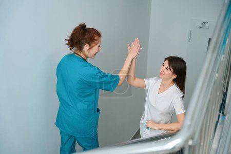 Téléchargez les photos : Heureuses femmes médecins en uniforme souriant et donnant cinq ensemble dans les escaliers à l'hôpital - en image libre de droit