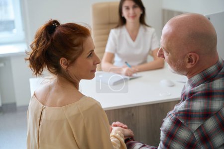 Photo for Lovely couple sitting with family practitioner at table and talking about IVF procedure in medical cabinet - Royalty Free Image