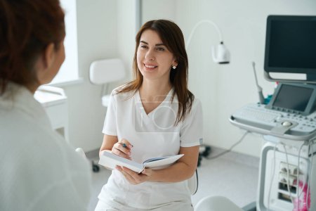 Téléchargez les photos : Thérapeute féminine joyeuse à l'écoute du patient et prendre des notes après le check-up à la clinique - en image libre de droit