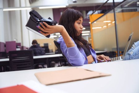 Téléchargez les photos : Belle femme avec ordinateur portable sur ses genoux travaillant tout en tenant un casque VR et assis dans un bureau moderne - en image libre de droit