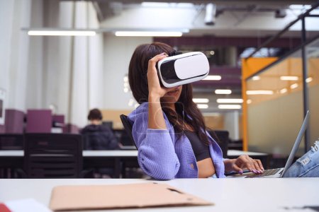 Téléchargez les photos : Attrayant femelle avec ordinateur portable sur ses genoux en utilisant un casque VR dans un bureau à aire ouverte - en image libre de droit