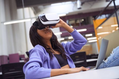 Téléchargez les photos : Belle femme avec ordinateur portable sur les genoux portant des lunettes de réalité virtuelle dans le bureau moderne - en image libre de droit
