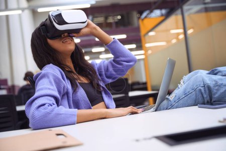Téléchargez les photos : Belle femme utilisant un casque VR tout en effectuant des tâches de travail sur ordinateur portable dans un bureau ouvert - en image libre de droit