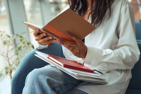Téléchargez les photos : Vue recadrée de la femme brune lisant les notes du carnet et assise sur un canapé confortable dans une chambre moderne - en image libre de droit