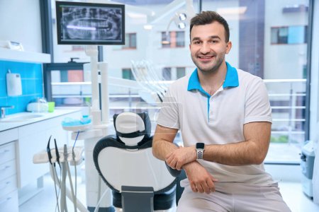 Téléchargez les photos : Homme souriant assis sur le lieu de travail dans le cabinet dentaire, le médecin a une montre intelligente - en image libre de droit
